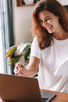 A cheerful woman in a white t-shirt working on her laptop, smiling warmly.