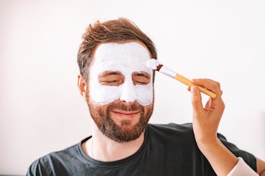 A man with a beard enjoys a spa treatment at home with a face mask applied with a brush.