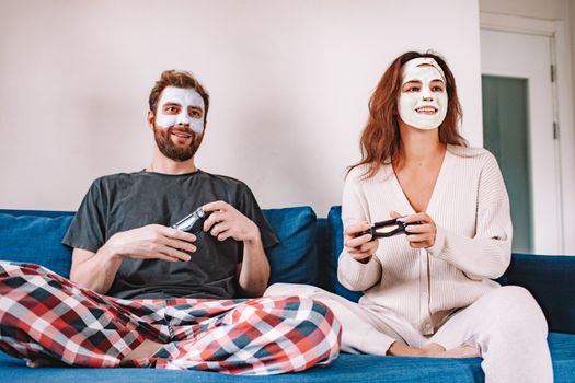 Happy couple with facial masks playing video games on the sofa, enjoying leisure time together.