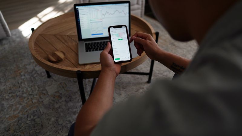 Man trading stocks online using smartphone and laptop. Indoor setting, focus on technology and finance.