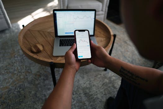 Person with tattoo reviews stock data on phone and laptop, indoors setting.