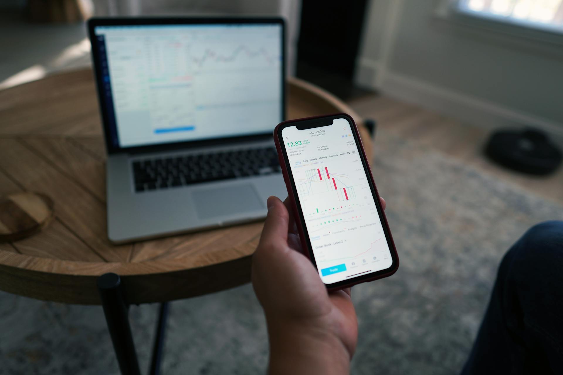 Person using smartphone with stock market charts while laptop displays data. Indoor setting.