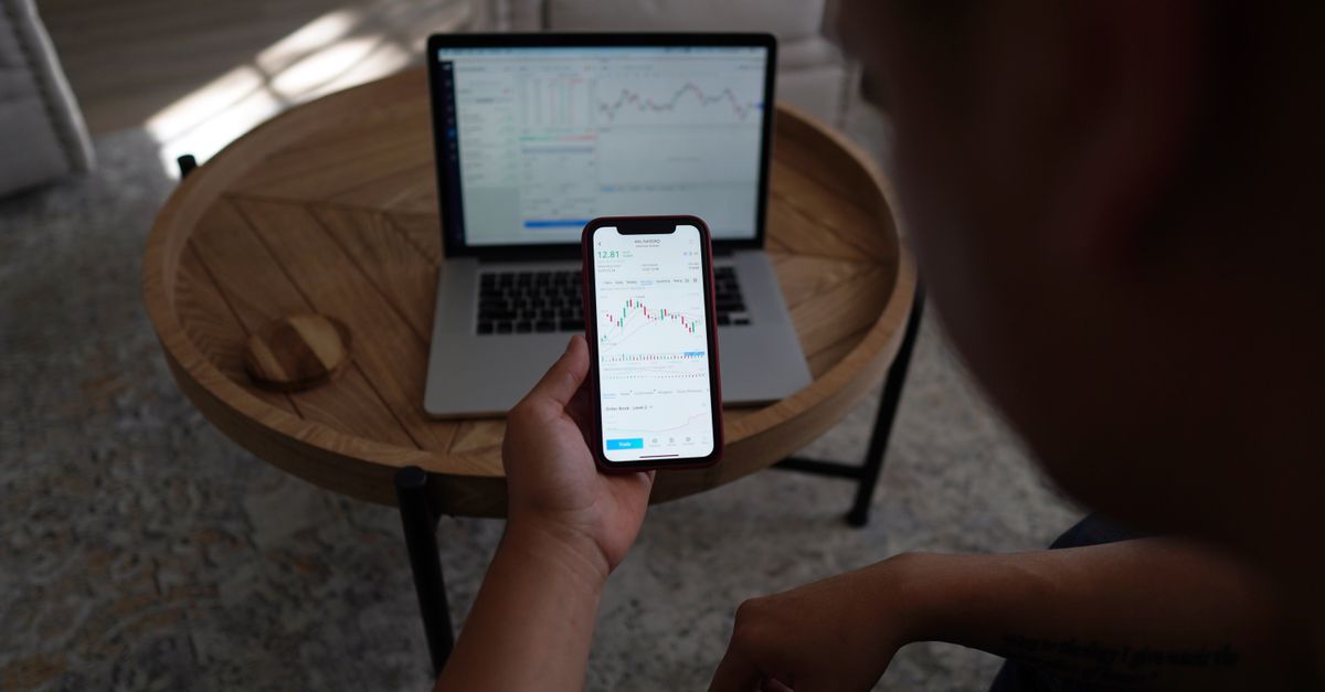 A young person holding a smartphone with stock market graph on screen, sitting in a cafe