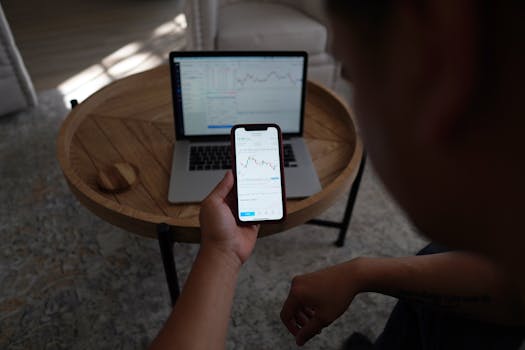A person examines stock market graphs on a smartphone and laptop at home, showcasing remote financial analysis.