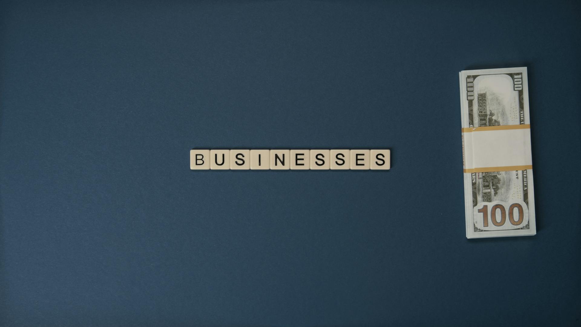 Dollar bills and 'BUSINESSES' spelled with tiles on a blue background, symbolizing finance.