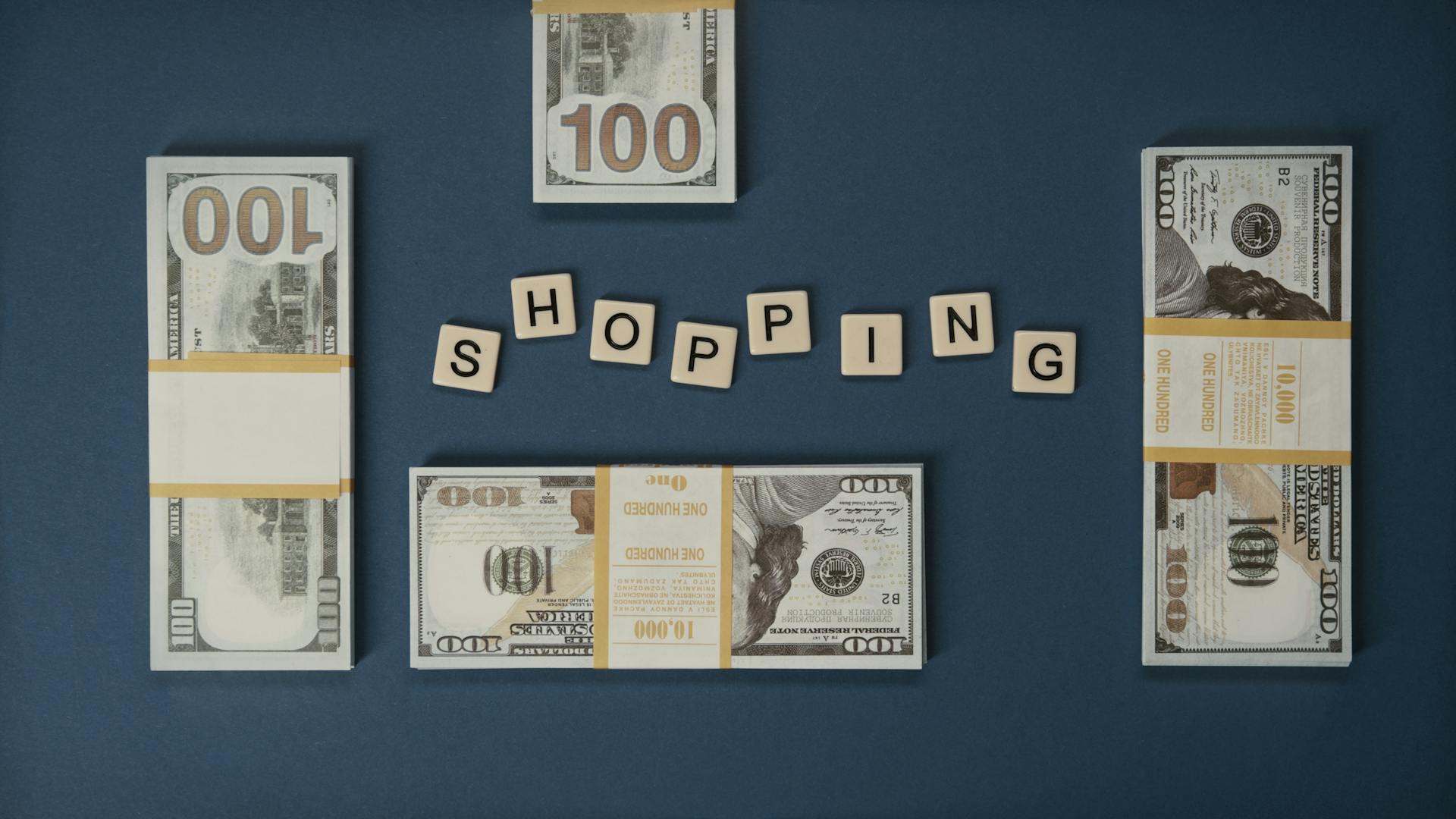 Flat lay of US hundred dollar bills and Scrabble tiles spelling 'shopping', evoking financial and retail themes.