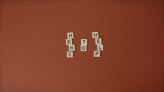 Flat lay of Scrabble tiles spelling 'Here to Help' on a brown background with copy space.