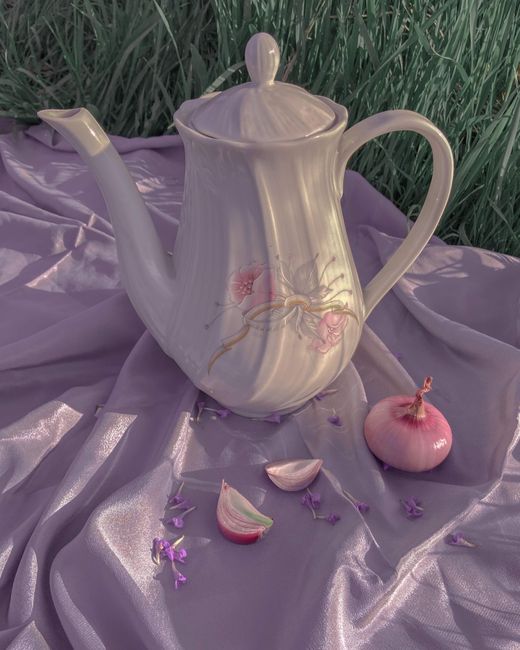 From above of porcelain teapot with small buds of flowers placed near onion on fabric in green meadow