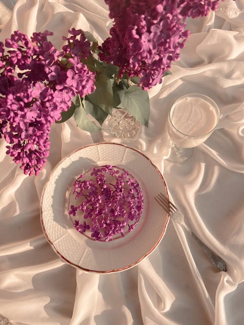 Top view of arranged bouquet of fresh flowers of lilac with plate of frozen flowers and fork near goblet of milk