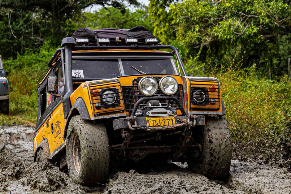 Rugged orange Land Rover navigating a muddy off-road track in dense forest terrain.