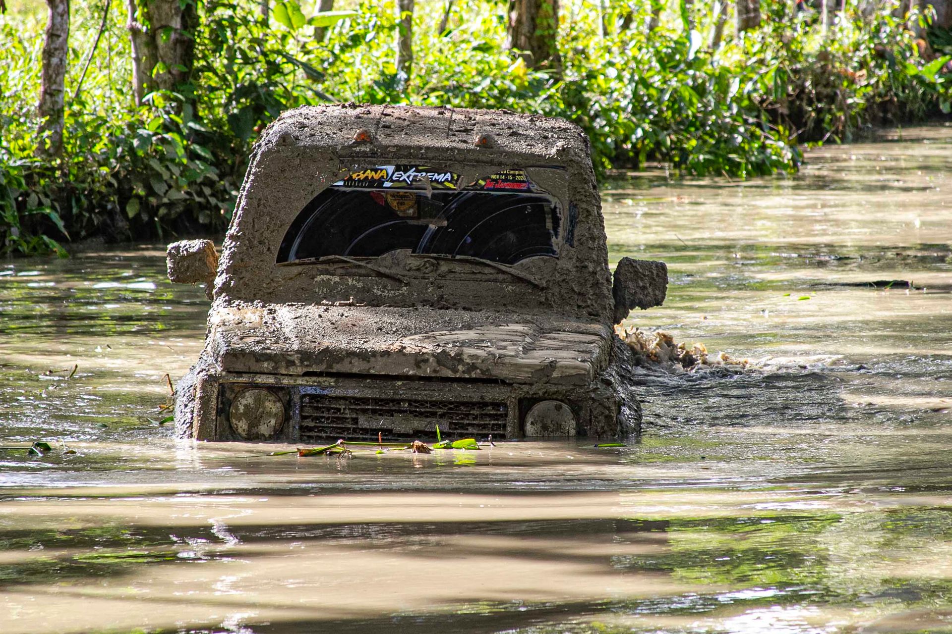 A muddy off-road vehicle submerged in water on a forest trail, showcasing tough terrain navigation.