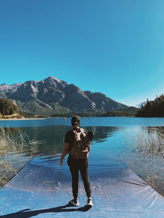 Full body back view of unrecognizable male hiker with backpack admiring rippling water of pond surrounded by mountains