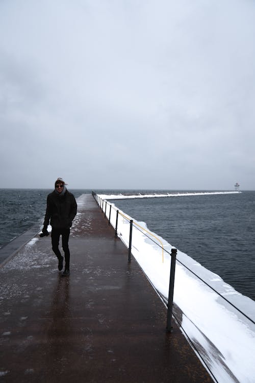 Full body of faceless man in warm clothes strolling along pear near deep sea in stormy weather in soft daylight