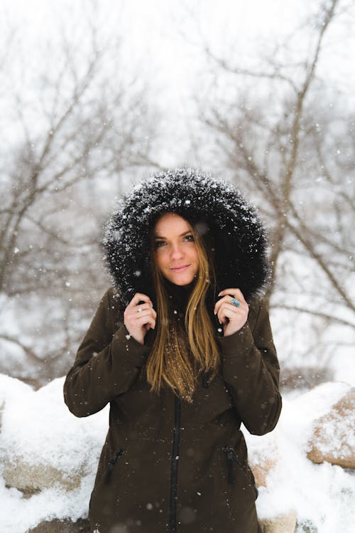 Cheerful young woman in warm clothes standing in winter forest and looking at camera in soft daylight