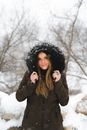 Cheerful young woman in warm clothes standing in winter forest and looking at camera in soft daylight