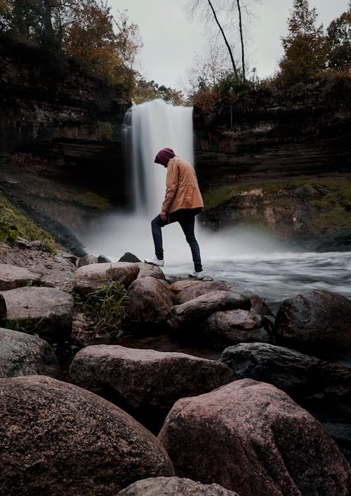 Side view of faceless man in warm clothes standing on rocky shore near waterfall in forest in evening in soft light