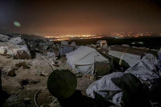 Refugee tents amidst rocky terrain with city lights at night in Idlib, Syria.