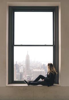 Woman sitting on a window sill with view of the Empire State Building in New York City. by Brady Knoll