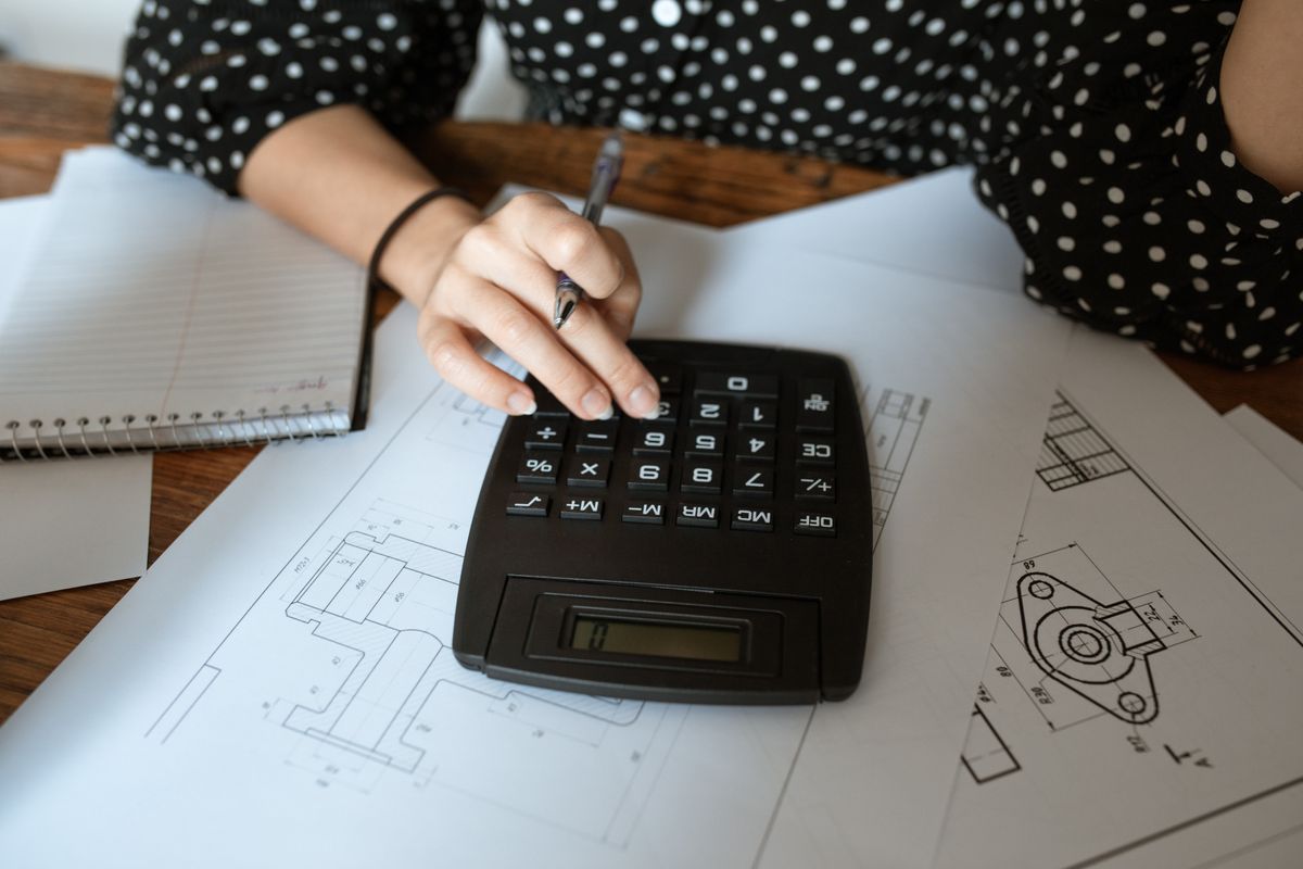 Masadaki teknik çizim kağıtlarının üzerinde siyah bir hesap makinesi var; bir el tuşlara basıyor, yanda defter bulunuyor.