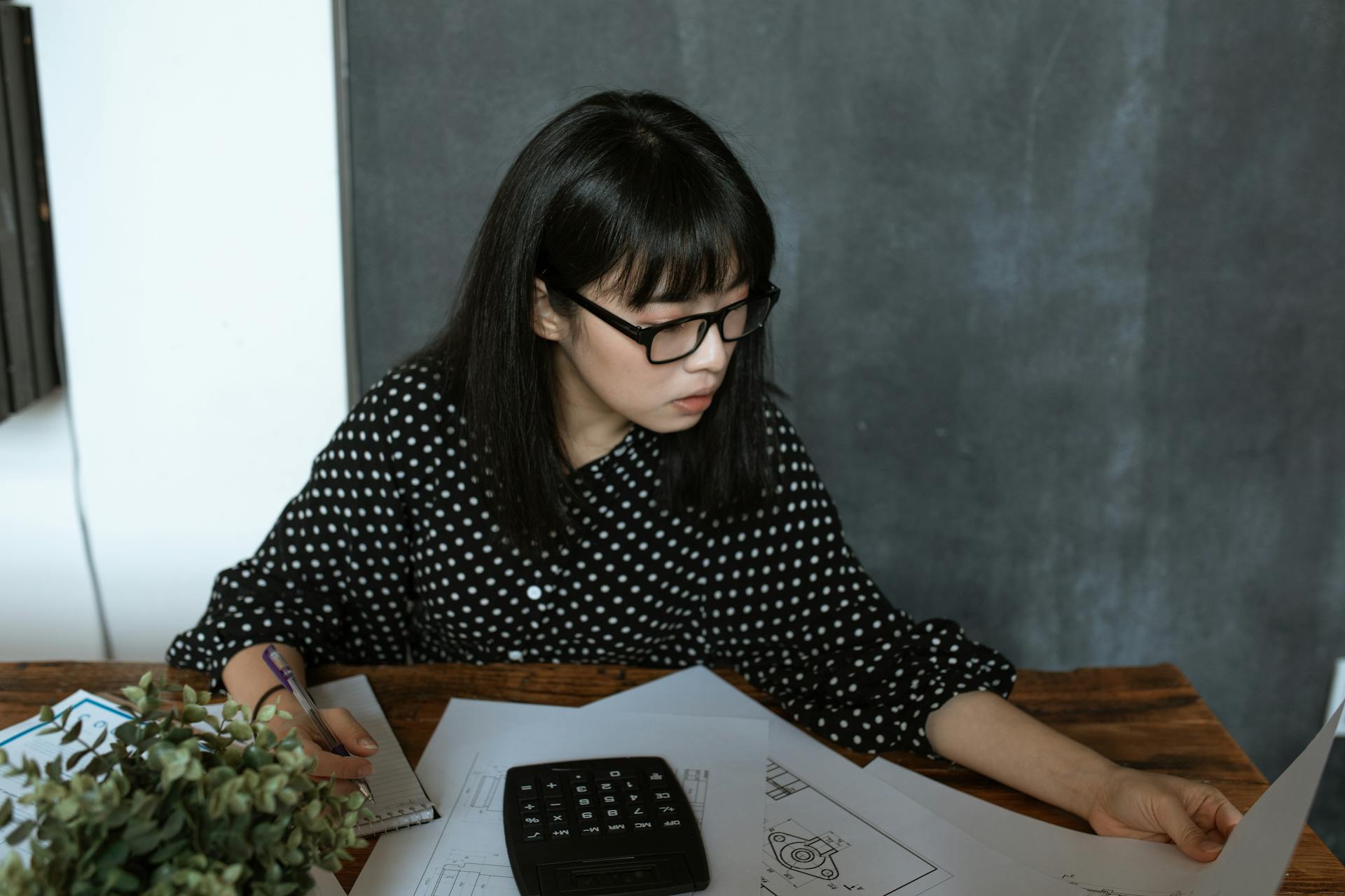 Asian businesswoman wearing glasses reviewing documents at her desk with a calculator nearby.