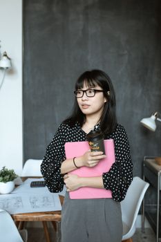 办公室里，一名穿着圆点衣服的年轻女子手拿咖啡杯和一本书站着