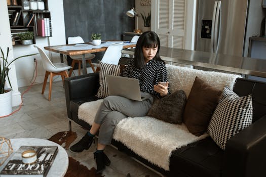 An Asian businesswoman working from home on a sofa, using a laptop and cellphone.