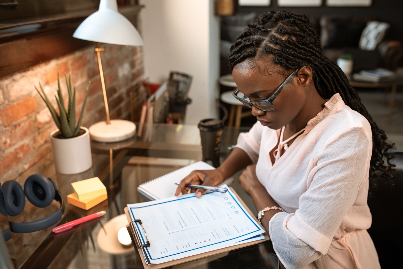 A Black woman in reading glasses points to a specific row on a clipboard form with checkbox bubbles and columns at a glass-topped home-office table, in a posture of careful side-by-side review.