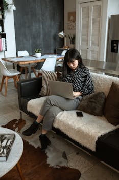 Asian woman sitting on a sofa, working on a laptop in a modern home interior setting.