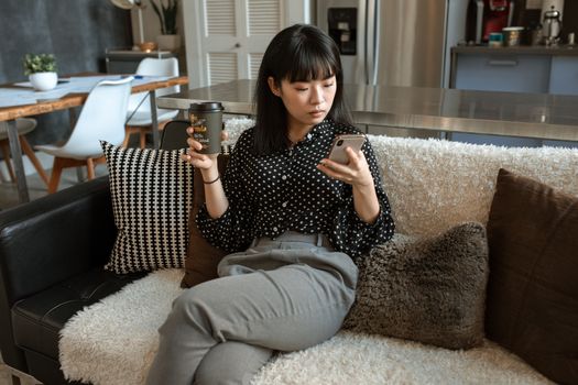 一名穿着圆点衬衫的年轻女子在舒适的客厅里边喝咖啡边用着智能手机