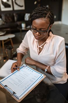 Woman with dreadlocks and eyeglasses reviewing documents in a home office setting. 5915211