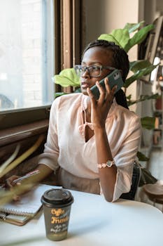 Young woman with eyeglasses talking on smartphone at cafe table with coffee, notebook, and plants.