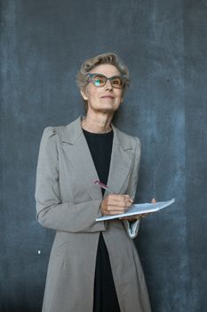 一位戴着眼镜、穿着灰色外套的老练女性，正微笑着在笔记本上写字