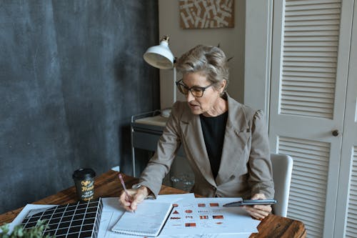 From above of concentrated senior female manager in formal outfit and eyeglasses taking notes in planner while working at table with various documents and takeaway coffee in cozy office