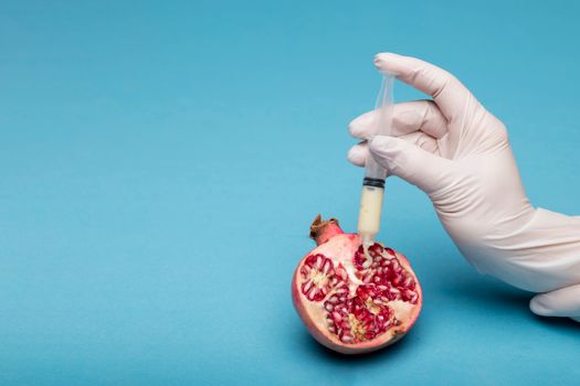 Gloved hand with syringe injecting into a pomegranate on a blue background.