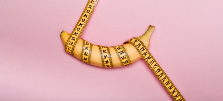 A banana wrapped with measuring tape on a pink background representing measurement or size concept.