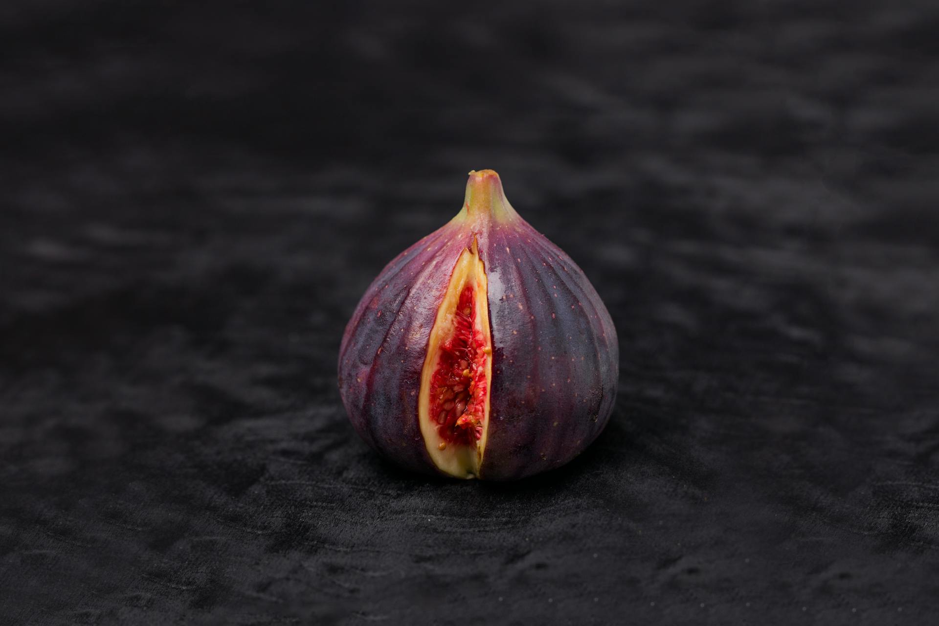 A close-up image of a single ripe fig fruit, sliced to reveal its vibrant interior, set against a dark background.