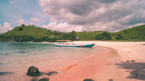 Free A serene beach with a boat on pristine pink sand, surrounded by lush green hills under a cloudy sky. Stock Photo
