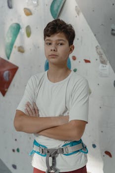 Teen boy standing confidently with crossed arms at indoor climbing gym, wearing a harness.