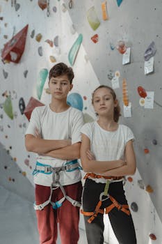Two teenagers in climbing gear, standing confidently in an indoor rock climbing gym.
