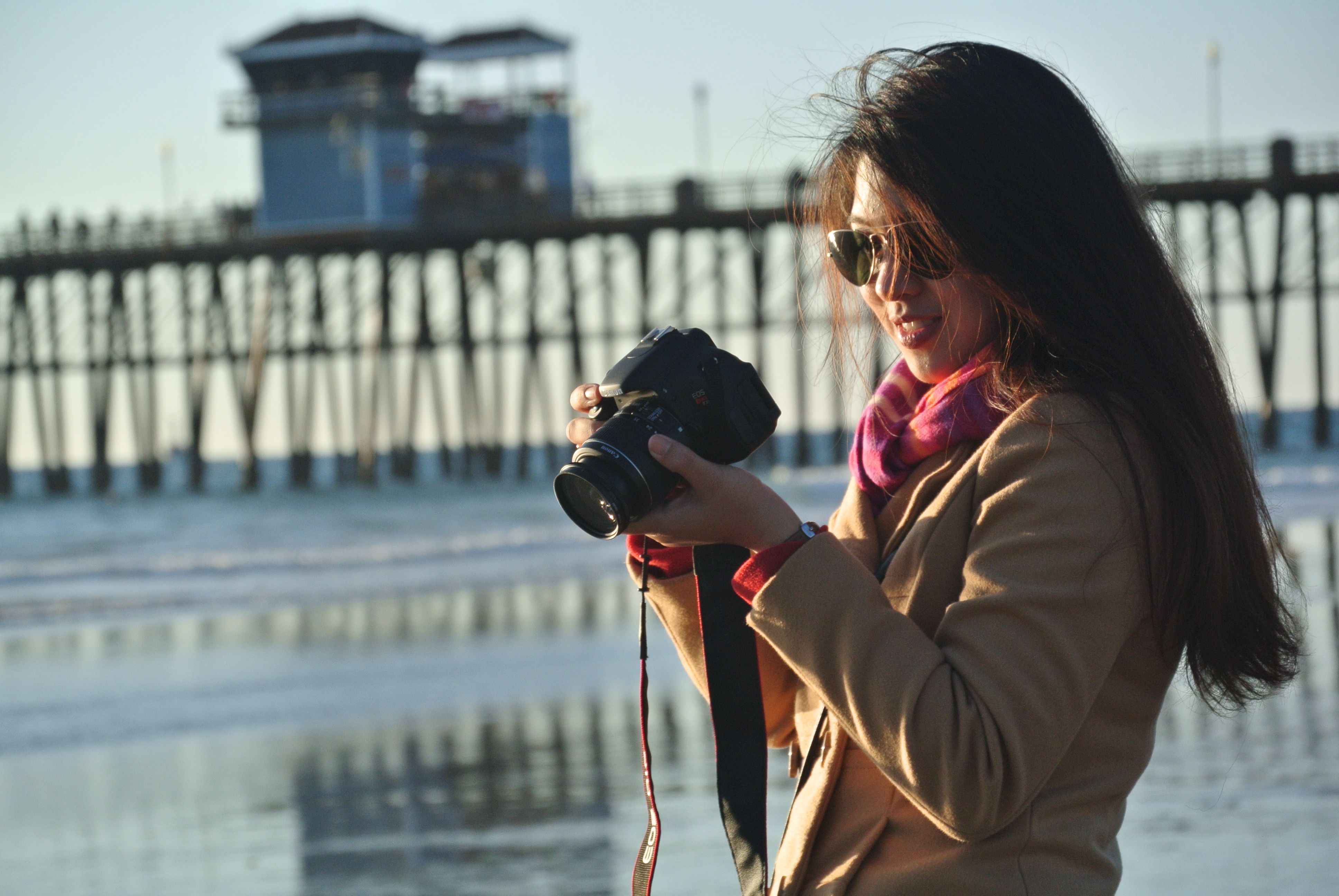 Smiling woman with sunglasses capturing photos by the beach pier.