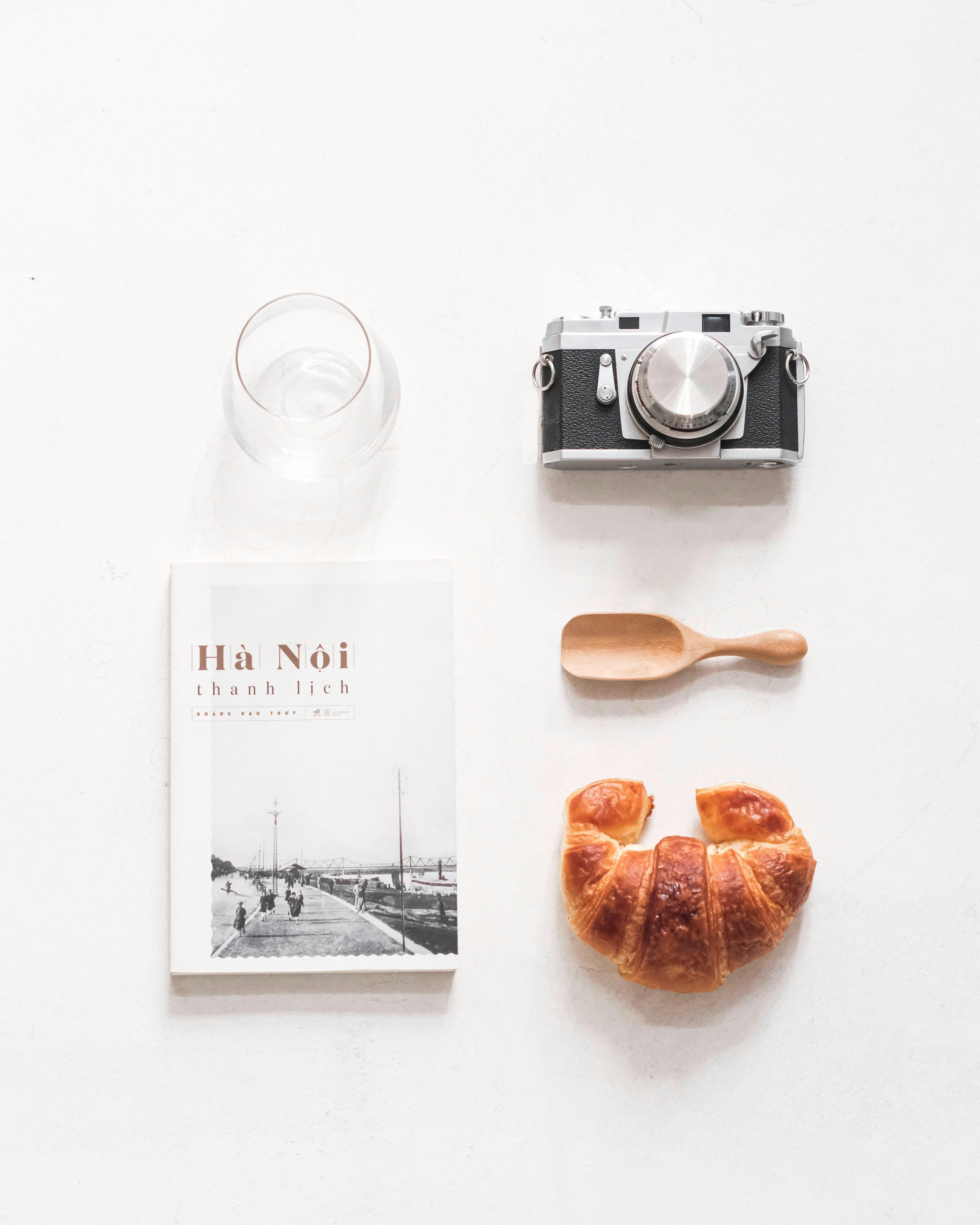 A minimalist top-down composition features a camera, book, croissant, glass, and spoon on a white background.