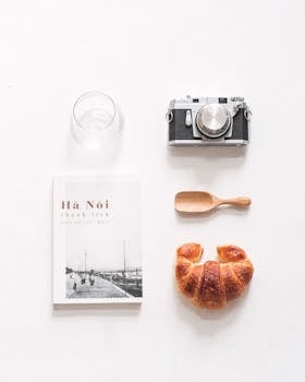 A minimalist top-down composition features a camera, book, croissant, glass, and spoon on a white background.