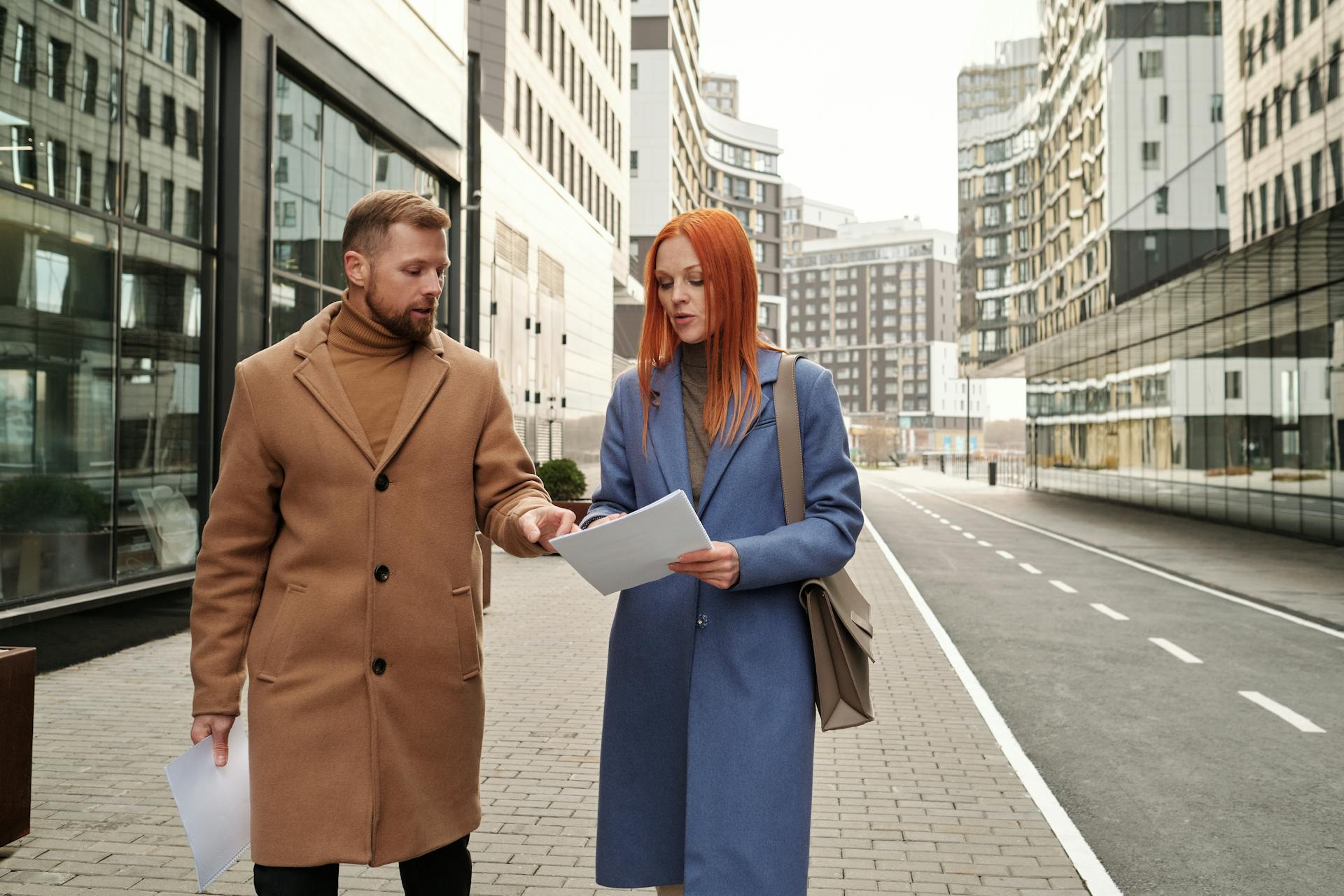 Two business professionals in winter attire reviewing documents in a modern cityscape.