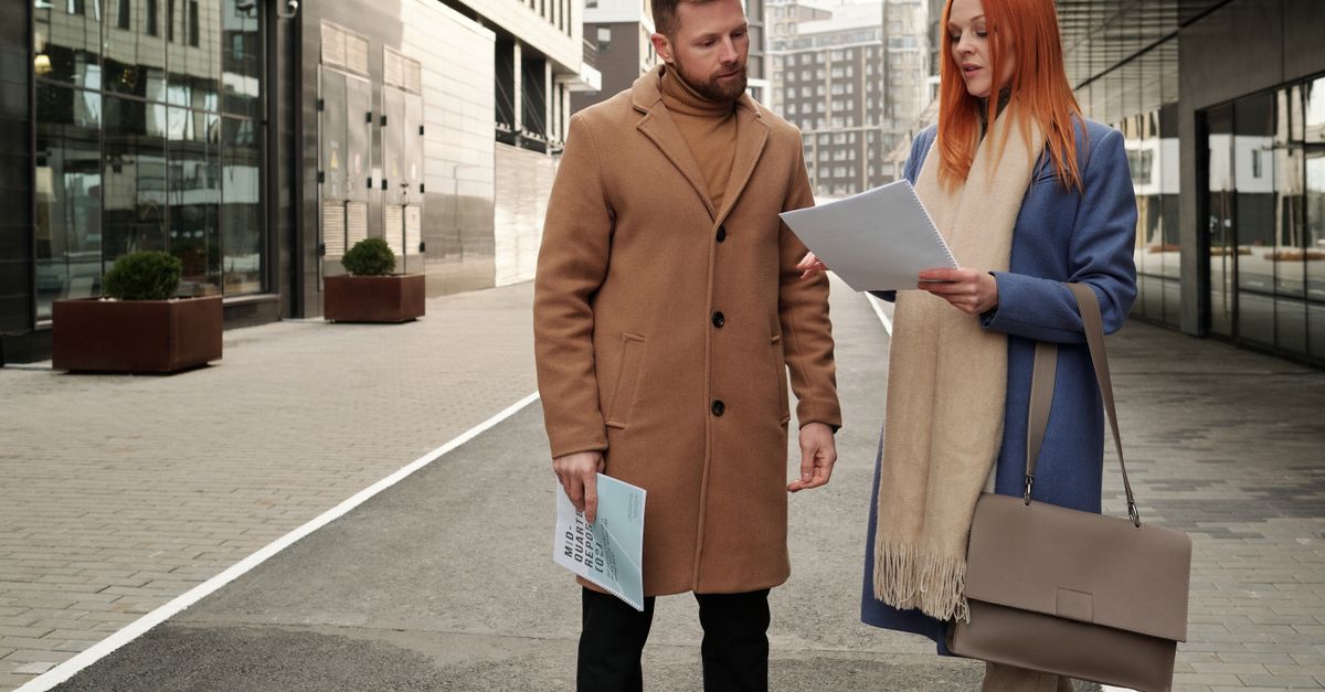 Two professionals walking outside while talking, one holding a notebook, with a cityscape in the background