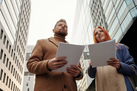Two professional individuals in corporate attire review documents in a bustling city environment.