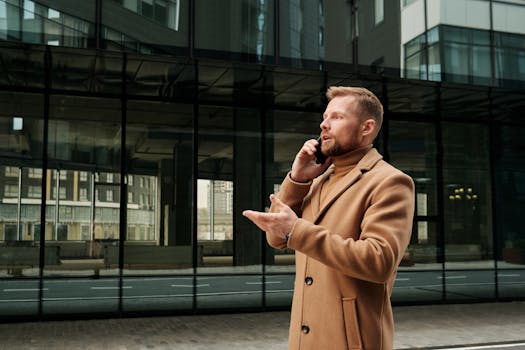 Confident businessman in a brown coat talks on phone outdoors in city.