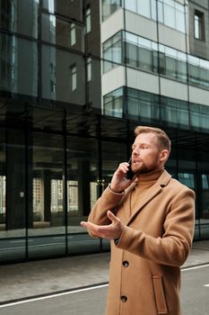 穿着棕色外套的自信商人在户外打电话，反对现代玻璃建筑