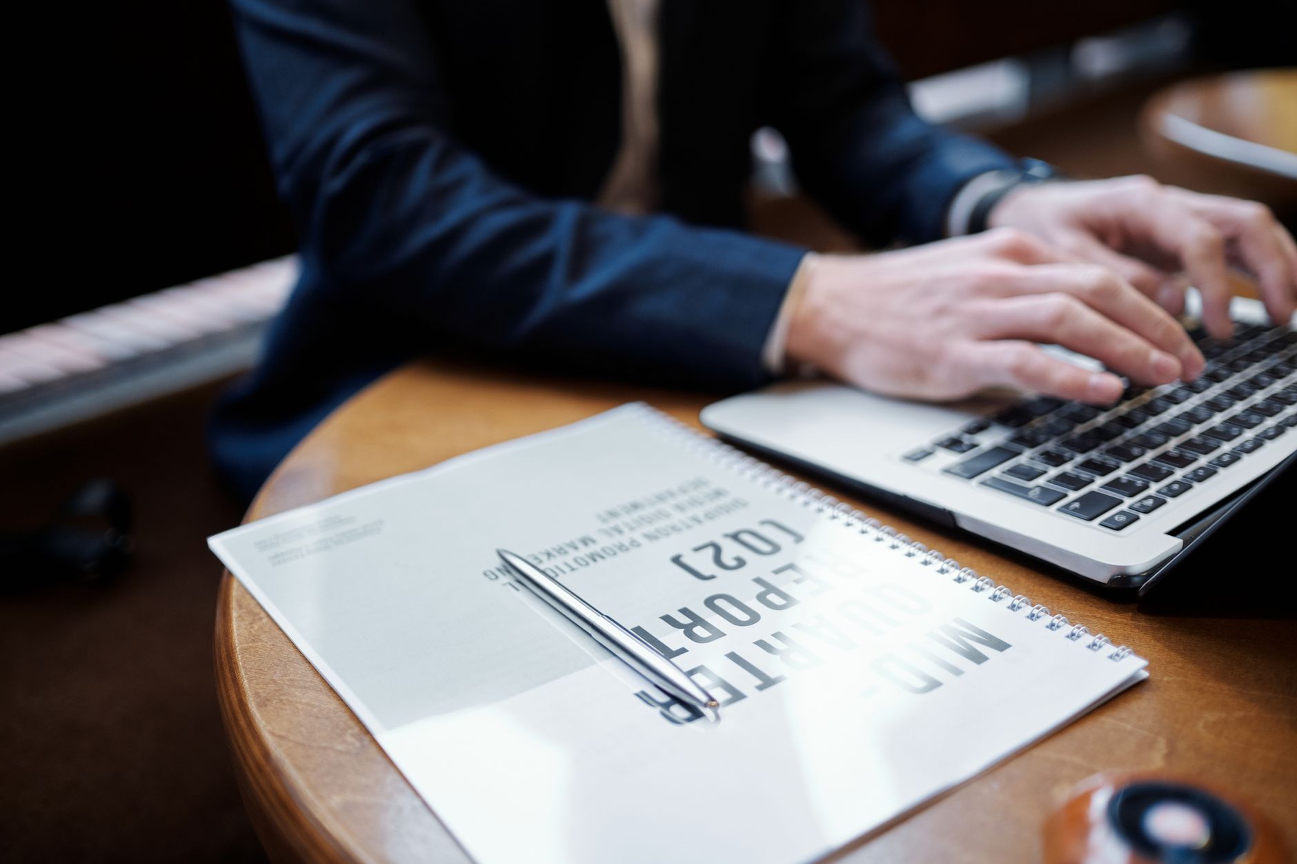 Businessperson typing on a laptop in an office setting with documents and pen.