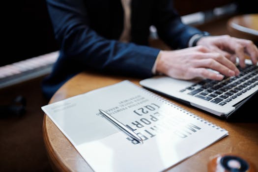 Businessperson typing on a laptop in an office setting with documents and pen.