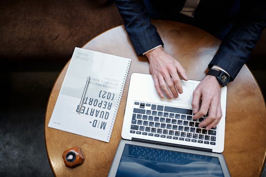 Top view of a businessman typing on a laptop next to a quarterly report on a wooden table.